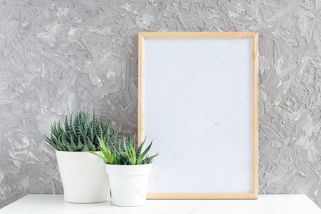 Wooden vertical white empty frame and two natural succulents flowers in white pots on table on gray concrete wall background. Mockup Template for your design