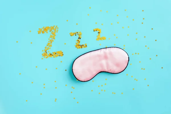 Pink sleep mask for eyes and gold stars confetti in form of dream symbols Z Z Z on blue background. Top view Copy space. Concept eye protection from light for good sleep and melatonin production