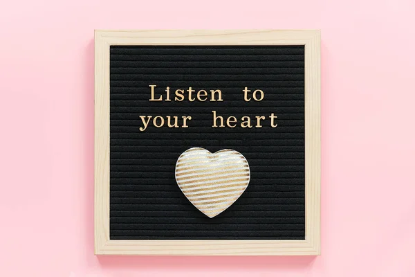 Listen to your heart. Motivational quote in gold letters and decorative textile heart on black letter board on pink background. Top view Flat lay Concept inspirational quote of the day