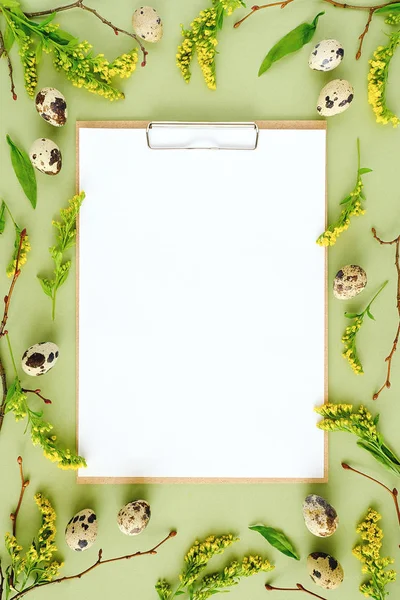 Spring easter floral frame and white blank paper. Natural tree branches, yellow flowers, quail eggs, clipboard notepad on green background Mockup Creative Flat lay Top view