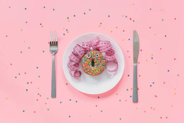 Abstracto rostro divertido de la mujer hizo donut con los ojos y el pelo de cinta de centímetro en el plato, cubiertos sobre fondo rosa. Comida rápida, engorde y creatividad alimentaria poco saludable Vista superior — Foto de Stock