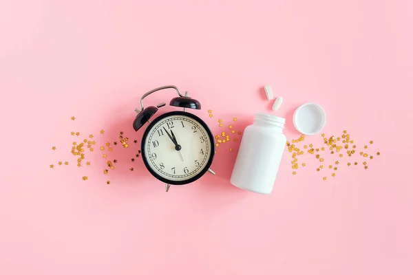 Zwei Pillen, weiße Flasche, schwarzer Wecker und Goldsterne-Konfetti auf rosa Hintergrund. Konzept Schlaflosigkeit, Schlafprobleme, Zeit, Tabletten zu nehmen. Draufsicht Flache Lagen Kopierraum — Stockfoto