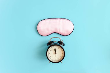Gözler için pembe uyku maskesi ve mavi arka planda siyah alarm saati. Gece yarısı oldu. Üst görünüm kopyalama alanı. İyi bir uyku ve melatonin üretimi için ışık koruması kavramı.