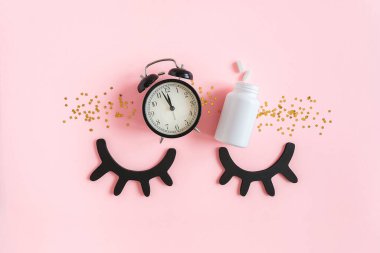 Kapalı dekoratif gözler, iki hap, beyaz şişe, siyah çalar saat ve pembe arka planda altın yıldızlı konfeti. Insomnia kavramı, uyku sorunları, ilaç alma zamanı. Üst Görünüm Düz yatay Boşluğu Kopyala.
