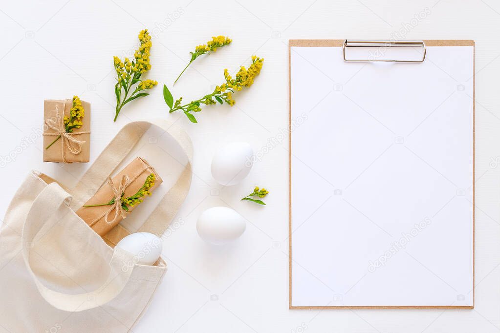 Happy easter concept. White eggs, yellow flowers, craft gifts fly out textile bag and clipboard with paper on white background Mockup Flat lay Top view.