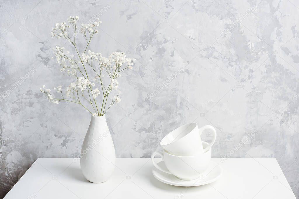 Bouquet of small delicate flowers gypsophila in vase and and two white cups for tea or coffee on table agains gray concrete wall. Copy space Front view.