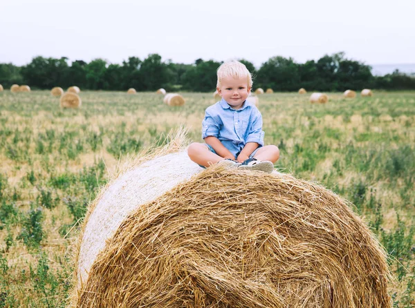 Χαριτωμένο μικρό παιδί αγόρι καθιστός σε Θημωνιές στο πεδίο σιτάρι. — Φωτογραφία Αρχείου