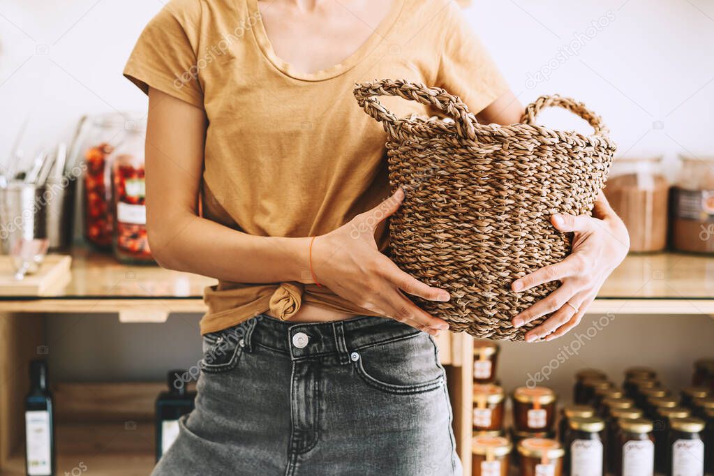 Woman with wicker basket, reusable cotton bag and glass jars buying in plastic free grocery store. Sustainable shopping in zero waste shop. Minimalist eco-friendly low waste lifestyle.