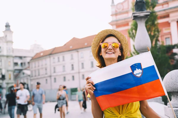 Young smiling girl in sunglasses with slovenian flag on central square of Ljubljana. Woman tourist holding slovenian flag on background of city architecture. Travel, living, study in Slovenia, Europe.