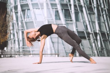 Güzel bir kadın şehir dışında yoga egzersizleri yapıyor. Modern gökdelenlerin arka planında yoga yapan genç bir kız. Sağlıklı yaşam tarzı, spor, şehir hayatı kavramı.