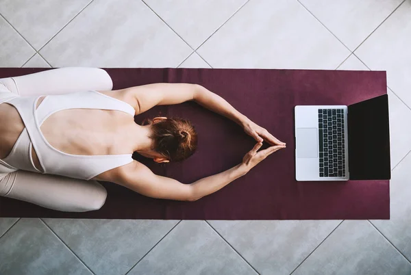 Top view of woman practicing yoga on yoga mat with laptop. Girl is meditating and relaxing with video training at home. Concept of online sport class or meditation course on digital devices.