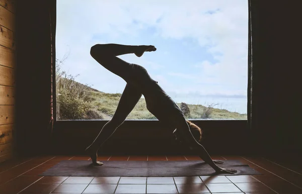 Silhouette of young woman practicing yoga indoor on background of big window with natural landscape. Girl doing vinyasa yoga flow practice on yoga mat. Female health of body and mind, relax at home.
