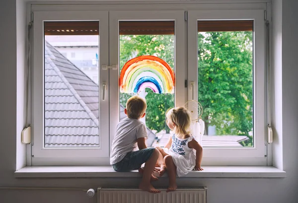 Cute little children on background of painting rainbow on window. Photo of kids leisure at home, safety joy symbol, happy childhood. Positive visual support during quarantine. Family Art Background