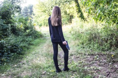 A Girl in black with a gun in the nature.  Agent. Long hair.