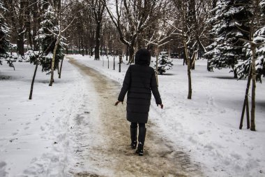 Siyah ceketli bir kız yol boyunca yürüyor. Bir kızın silueti. Kış yolu. Kışın kapüşonlu kız. Siyah kıyafetler. Ağaçlar ve karla dolu bir kış günü.