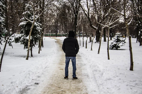 A guy in a black jacket is walking along the road. Silhouette of a man. Winter road. The man in the hood in the winter. Black clothes and jeans. A winter day with trees and snow.