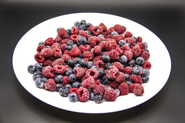 A Berry mix  from frozen raspberries and blueberries on the white plate. A Frozen Berries in black background.  A sweet background with frozen raspberries and blueberries