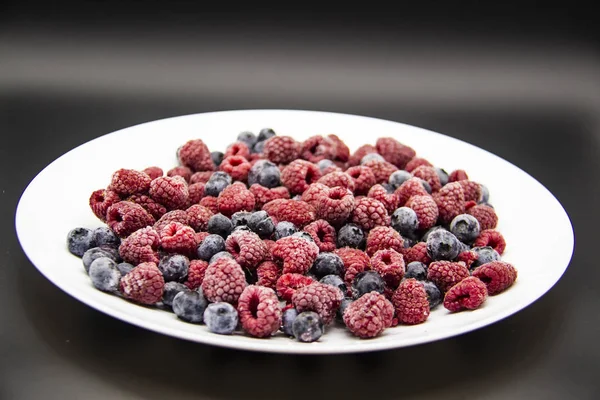 A Berry mix  from frozen raspberries and blueberries on the white plate. A Frozen Berries in black background.  A sweet background with frozen raspberries and blueberries
