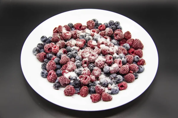 A Berry mix in sugar from frozen raspberries and blueberries on the white plate. A Frozen Berries with Sugar.  A sweet background with frozen raspberries and blueberries