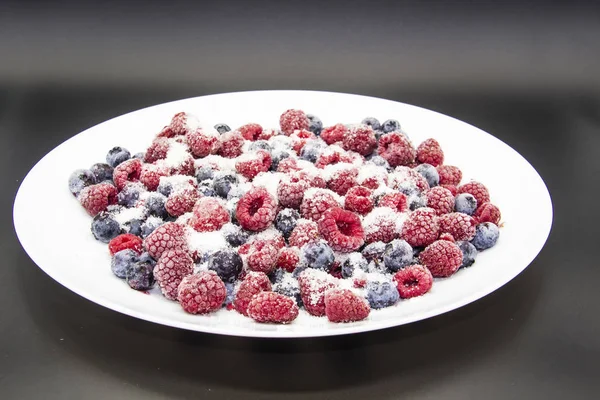 A Berry mix in sugar from frozen raspberries and blueberries on the white plate. A Frozen Berries with Sugar.  A sweet background with frozen raspberries and blueberries