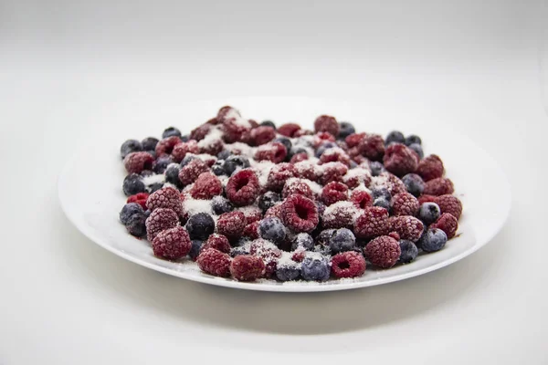 A Berry mix in sugar from frozen raspberries and blueberries on the white plate. A Frozen Berries with Sugar.  A sweet background with frozen raspberries and blueberries