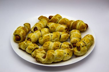 Beyaz arka planda beyaz bir tabakta hamurda pişmiş sosisler. Sosisli börek. Sosisli börek. Puf böreğe sarılmış ve tabakta pişirilmiş. Lezzetli sosisli mini çörekler..