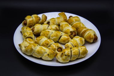 Beyaz bir tabakta siyah arka planda hamurda pişmiş nevaleler. Sosisli börek. Sosisli börek. Puf böreğe sarılmış ve tabakta pişirilmiş. Lezzetli sosisli mini çörekler..