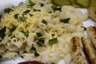 Üstünde peynir ve yeşil soğan olan bir tabakta makarna. Izgaradan tavuk sosisi ve kavanozdan dilimlenmiş salatalıkla yüksek kaliteli undan yapılmış lezzetli bir makarna. Peynir, maydanoz, sosis, soğan ve sosla pişirilmiş un boynuzları.