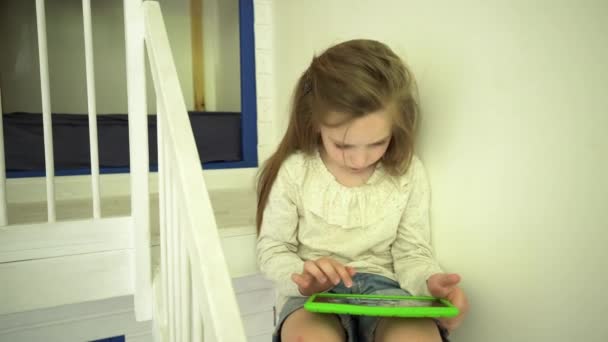 Komoly aranyos kislány ül az ágy lépcsőjén táblagéppel. Kézi — Stock videók