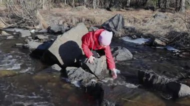 Kız bir kayanın üzerinde oturuyor ve soğuk bir akıntıyla oynuyor, parmakları soğuk ve soğuk sudan soğuk.