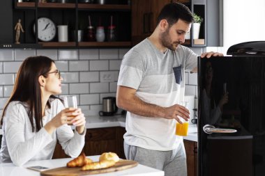 Couple in the kitchen eat breakfast with orange juice and pastry at table and he open refrigerator