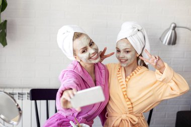 Anne ve kızı telefonda yüzlerinde maske ve kafalarında havluyla selfie çekiyorlar.