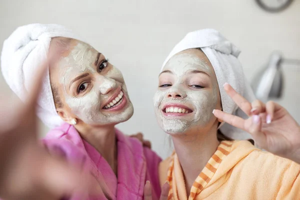 Mother and her daughter taking selfie by phone with facial mask and towel on head