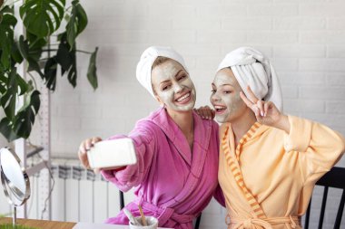 Anne ve kızı telefonda yüzlerinde maske ve kafalarında havluyla selfie çekiyorlar.