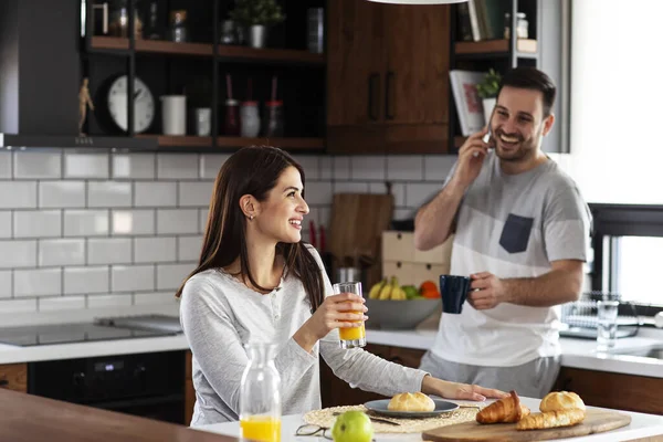 Couple in the kitchen eat breakfast with juice and pastry on table and he is talking on the mobile phone while she drink orange juice