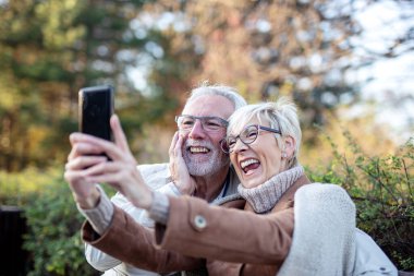 Yetişkin bir çift parktaki bankta otururken cep telefonuyla selfie çekiyor.