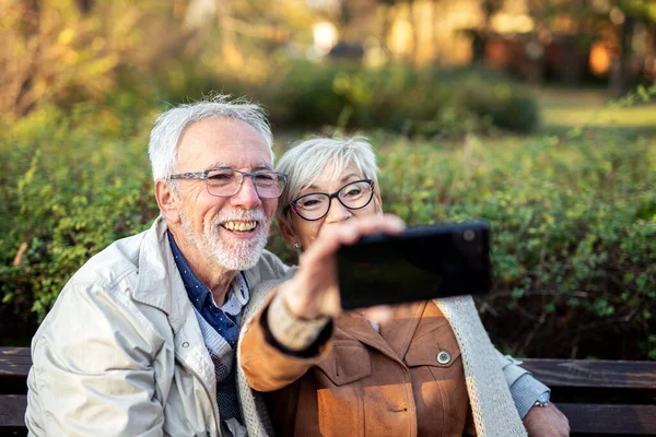 Yetişkin bir çift parktaki bankta otururken cep telefonuyla selfie çekiyor.