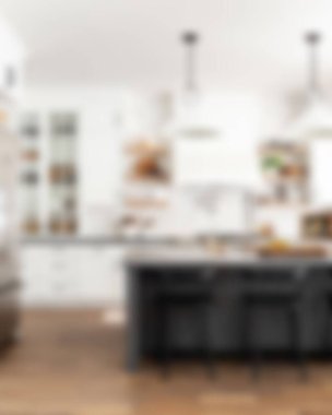 Ada, granit tezgah, paslanmaz çelik buzdolabı, fırın ve ahşap zemini olan çağdaş beyaz bir mutfağın içi.