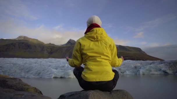 En flicka i en klargul jacka mediterar i en yogaposition på en sten framför en enorm glaciär — Stockvideo