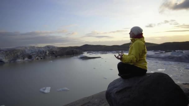Una chica con una chaqueta de color amarillo brillante medita en una pose de yoga sobre una piedra frente a un enorme glaciar. Relajación con vistas a las montañas y acantilados — Vídeos de Stock