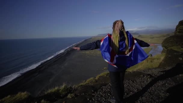 İzlanda bayrağı taşıyan güneş gözlüklü bir kız uçurumun kenarında duruyor. — Stok video