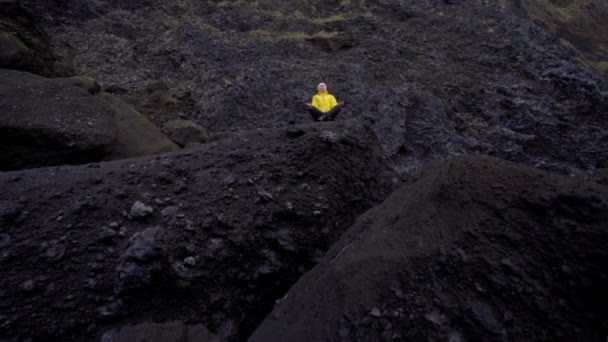 Une fille vêtue d'une veste jaune vif médite dans une pose de yoga sur un rocher noir. Détente près de l'océan — Video