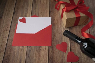 Sevgililer Günü için arka plan. Bir şişe şarap, kraft kağıdından bir hediye, kırmızı bir zarf, bir kalp ve ahşap arka planda saten kırmızı bir kurdele.