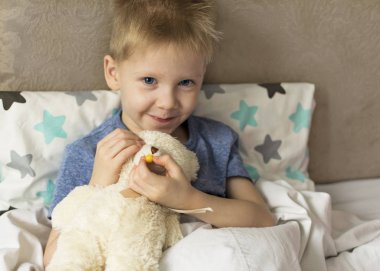 Ateşli ve başı ağrıyan hasta üzgün bir çocuk oyuncak bir ayıyla yatakta yatıyor. Grip salgını virüsü bakterisi