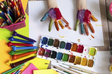 Çocuk resim yapıyor. Çok renkli suluboya bir çocuğun avuç içi ve elleri. Suluboya, guaj, boya fırçaları, renkli boya kalemleri, çıkartmalar. Çizim, yaratıcılık ve hobiler için hazır.