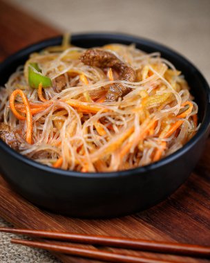 Glass noodle salad with meat and carrots in a black bowl on a wooden board