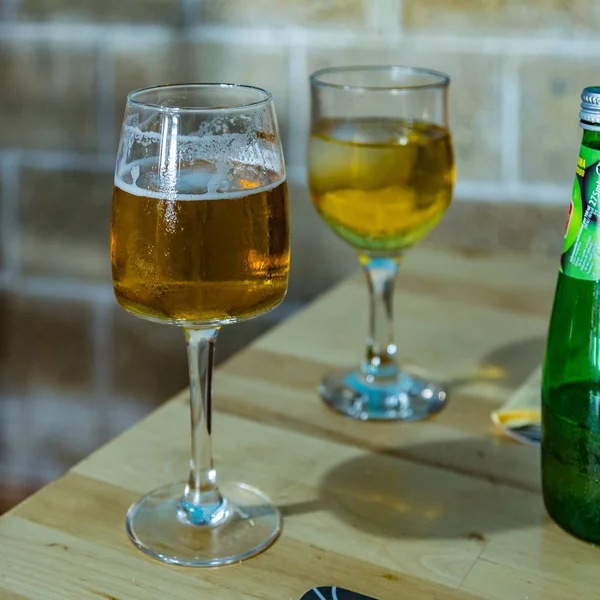 Sur la table à manger se trouve un verre de petite bière mousseuse et une bouteille verte. Gobelet froid de jus de pomme pétillant avec un glaçon sur fond flou. Focus sélectif, vue rapprochée. Cadre carré — Photo