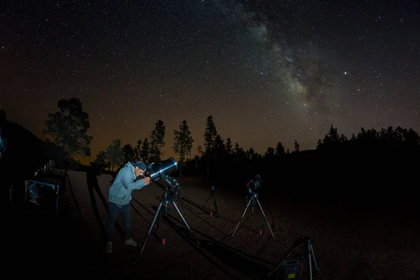 Unge man observerar stjärnhimlen genom ett teleskop. Berg, omgiven av tallskog i bakgrunden nattlandskap med färgglada Vintergatan Galaxy, stjärnor, planeter och fallande stjärna — Stockfoto