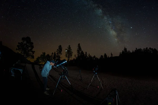 Молодой человек наблюдает звездное небо через телескоп. Горы, окруженные сосновым лесом на заднем плане ночной пейзаж с красочными Млечный Путь Галактика, звезды, планеты и падающая звезда — стоковое фото