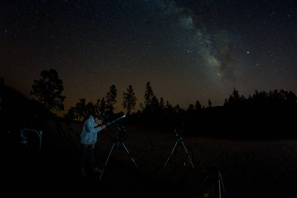 O jovem observa o céu estrelado através de um telescópio. Montanhas, rodeado por floresta de pinheiros na paisagem noturna de fundo com colorido Via Láctea Galáxia, estrelas, planetas e estrela cadente — Fotografia de Stock